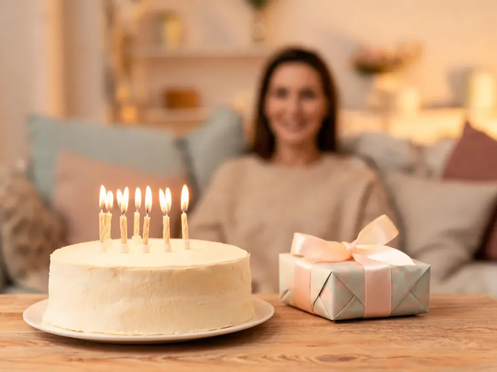 Elegancki tort z zapalonymi świeczkami i prezentem na stole, w tle uśmiechnięta kobieta tworząca ciepły, uroczysty nastrój.